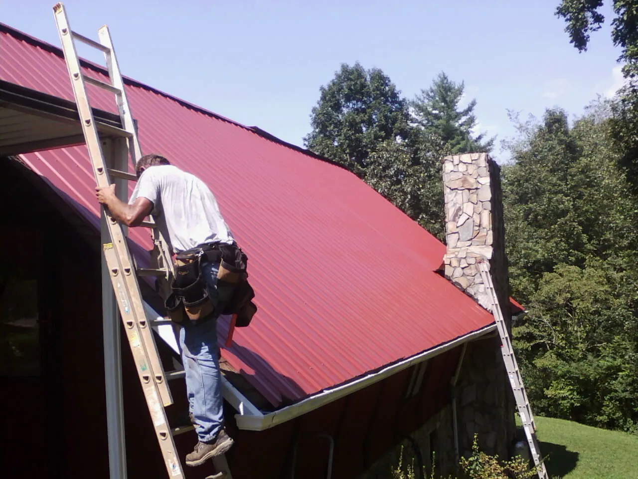 Roofer on a ladder beside a red metal roof and stone chimney — Soffit & Fascia Repair by Roofing Brothers in Brightwood